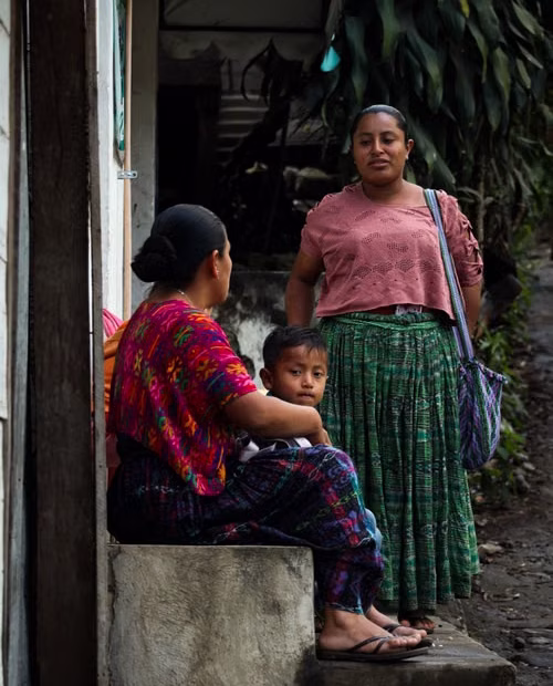 Señora indígena parada junto a una puerta y junto a ella una muchacha sentada y un niño pequeño parado junto a ella