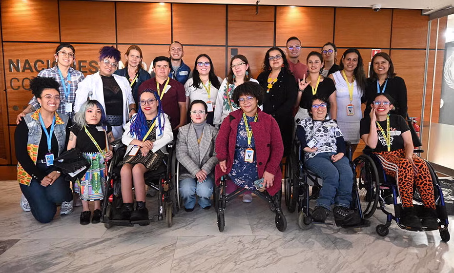 Grupo de mujeres con discapacidad , la mayoría usuarias de sillas de ruedas, en un escenario frente a varias personas en un salón